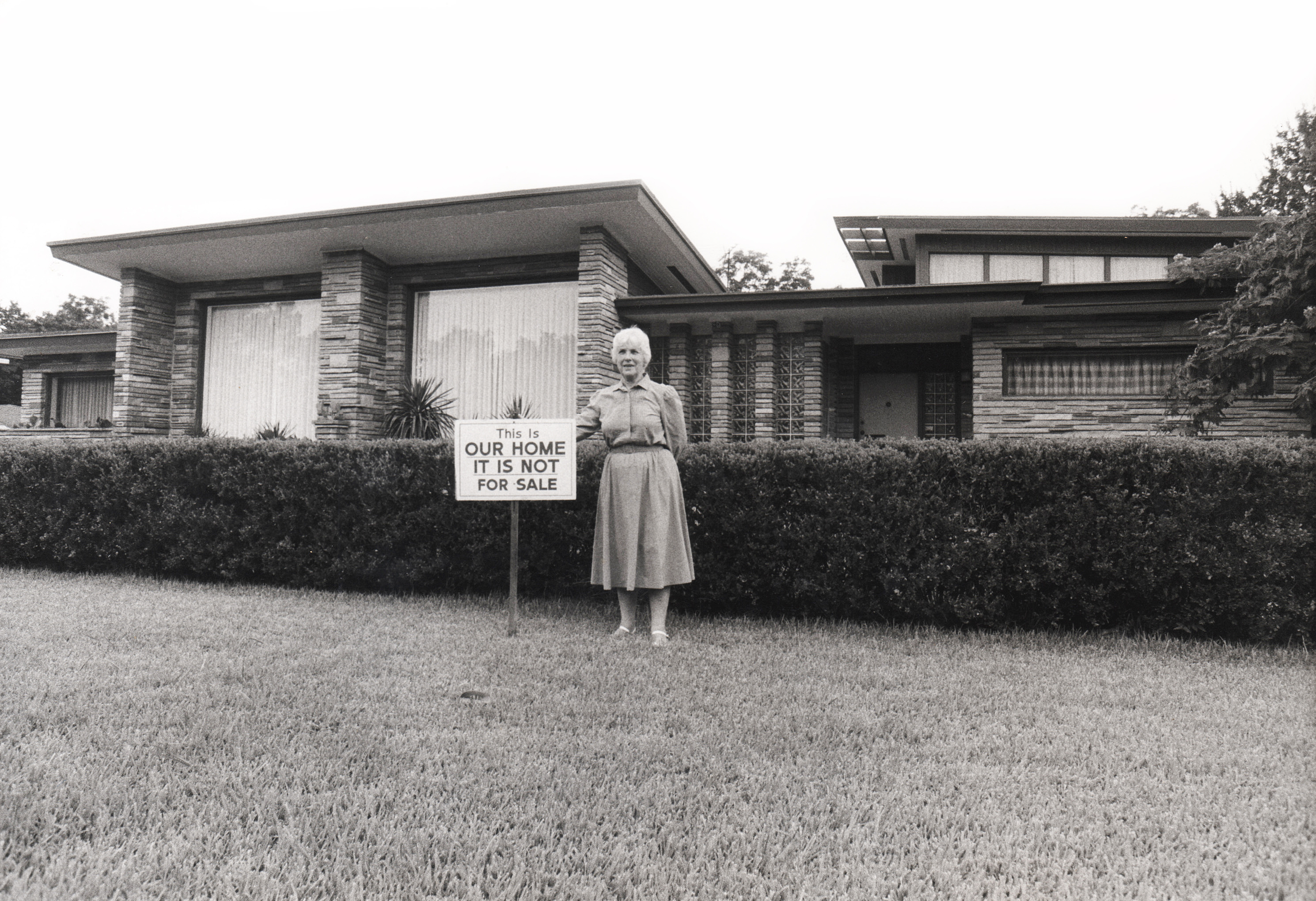 A white woman stands in front of a house next to a sign that reads: This Is OUR HOME IT IS NOT FOR SALE.